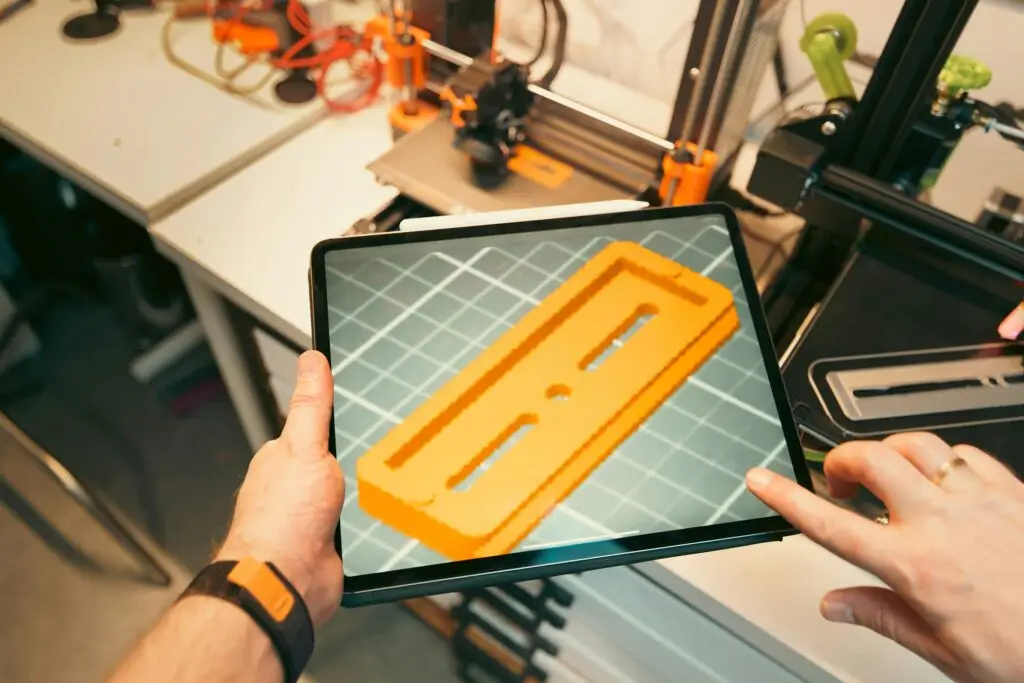 A person uses a tablet to view a 3D model prototype in a workshop equipped with 3D printers. Emkatech 3D-Druck Beratung – Konstruktion und Dokumentation